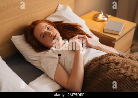 Top view of sick young woman with flu lying under blanket on bed holding thermometer in armpit, looking at camera. Ill female with cold lying in bed Stock Photo