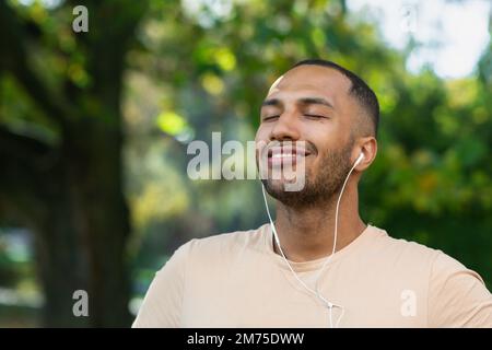 Close-up portrait of sportsman in park, hispanic man jogging in park with eyes closed breathing fresh air and resting, jogging with headphones listening to music and online radio and podcasts. Stock Photo