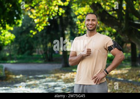 Portrait of successful man in park hispanic man with headphones listening to music and audiobooks online podcasts, smiling and looking at camera while jogging and doing fitness exercise. Stock Photo