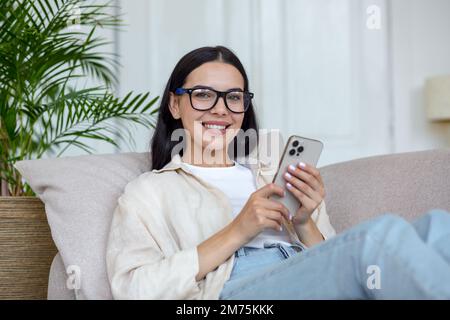 Young beautiful woman in glasses is lying on sofa and using mobile phone, looking at camera, smiling. Stock Photo