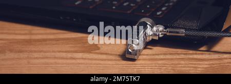The black laptop is locked with a cable lock. Panorama of laptop connectors lying on a wooden table. Stock Photo