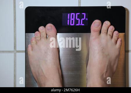 Stainless steel digital scale with mans feet weighing 185 pounds on tile floor Stock Photo