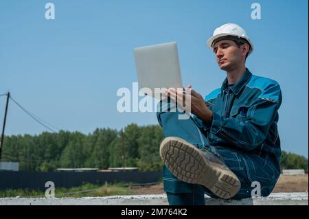 Caucasian male builder in hardhat sits on floor slabs and uses laptop at construction site.  Stock Photo
