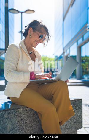 Happy attractive business woman middle-aged working smiling with laptop in front of a modern building. Vertical photo. Stock Photo