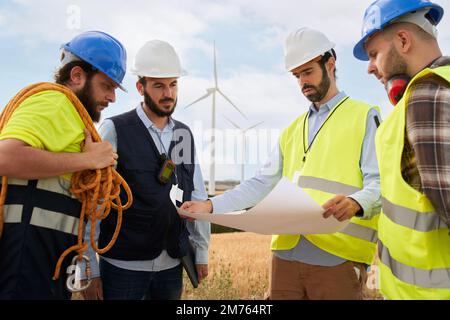 Group of engineers talking and working with a blueprint in an agricultural field with wind turbines Stock Photo