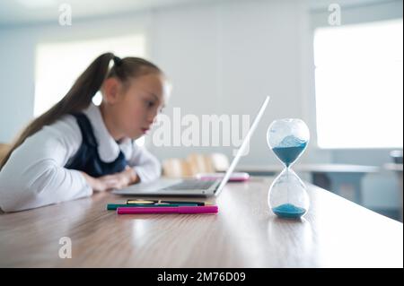 Caucasian girl sits at a desk at school and studies at a laptop. Hourglass on the table. Stock Photo