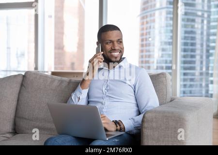 Handsome African man makes call sits on sofa with laptop Stock Photo