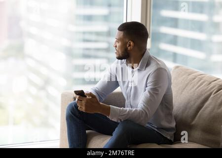 African man resting on couch holds smartphone staring out window Stock Photo