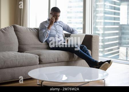 Man sits on couch working on laptop, talking on phone Stock Photo