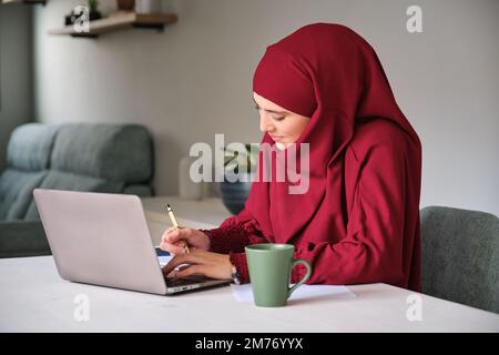 Muslim young woman student in hijab using laptop at home. Stock Photo