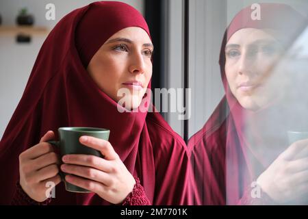 Muslim woman in hijab with a coffee cup looking through a window. Stock Photo