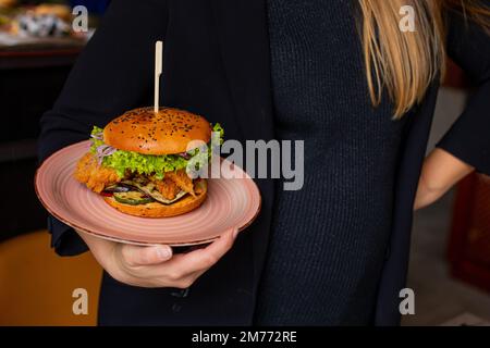 Woman in black clothes hold in hand plate with juicy appetizing chicken burger in public catering. Serving of big caloric sandwich on skewer. Like Stock Photo