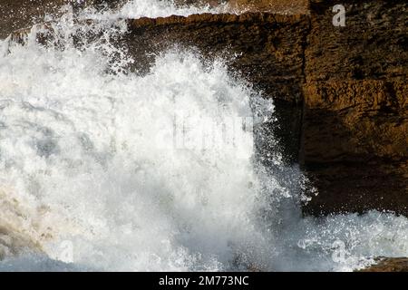 Atlantic Ocean waves crash into dramatic layered rock formations near Guincho Beach, Cascais, Portugal, under bright winter sunlight. Stock Photo