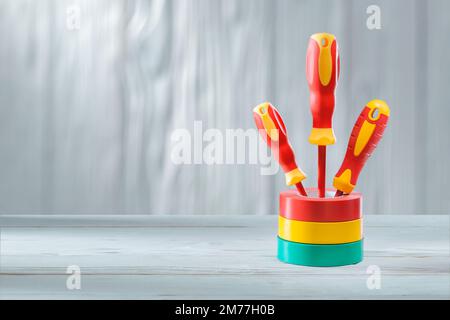 three red yellow insulated electrical screwdrivers standing in stack of rolls insulation tapes vintage white painted wooden background Stock Photo