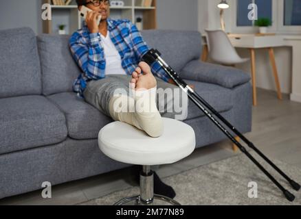 Young man with injured leg sitting at home on sofa with crutches and talking on the phone. Stock Photo