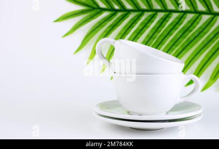 White mug and saucer on a white background, near palm leaves. A set of porcelain or ceramic dishes. Copy space Stock Photo