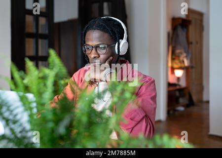 Thoughtful black freelancer man in earphones looking at laptop screen thinking solving work problem. Stock Photo