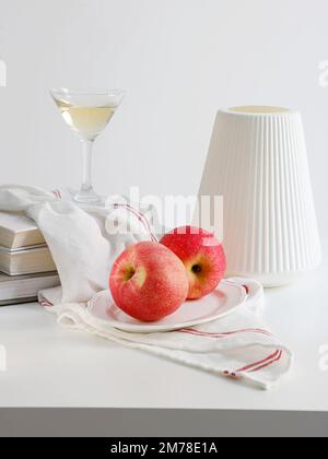 A vertical shot of a plate of red apples, a vase, a kitchen towel and a martini glass on white table Stock Photo