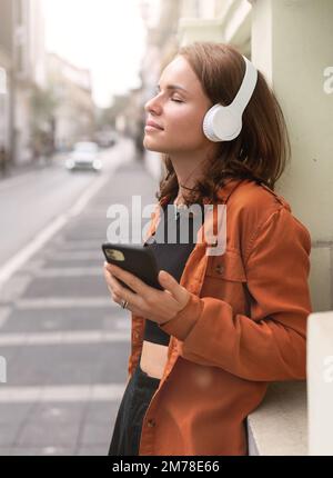 Young brunette woman standing on street with closed eyes listening to music with headphones and play list in her smart phone. Stock Photo