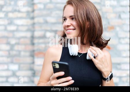 Attractive stylish young brown haired woman with big white headphones on her neck standing outdoors against brick wall holding mobile phone in hand an Stock Photo
