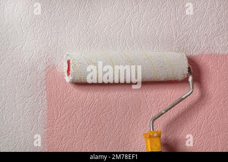 An old, fuzzy paint roller draws pink wallpaper on the wall with white paint. Copy space. Stock Photo
