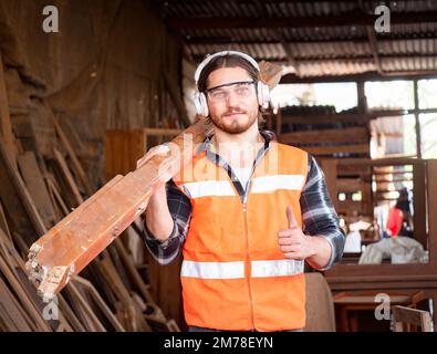 Portrait of handsome Caucasian carpenter carries a wood plank on his shoulder in the carpentry workshop. Preparing raw materials for the future woodwo Stock Photo