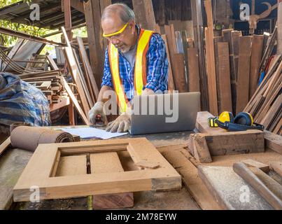 Happy senior carpenter using a tape measure for measuring woodwork in the carpentry workshop. Manufacture of wood products. Stock Photo