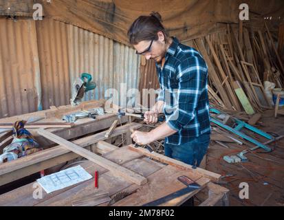 Handsome Caucasian carpenter craftsman wearing safety gear working with hardwood using a miter saw to cut a piece of wood in the workshop. Manufacture Stock Photo