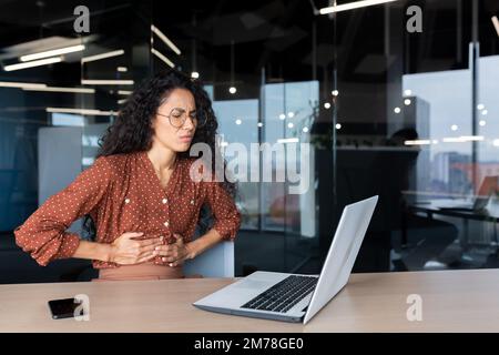 Abdominal pain at the workplace. A young Latin American woman is sitting at the desk in the office, holding her stomach with her hands, grimacing in pain. Needs help, treatment. Stock Photo