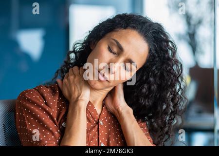 Close-up photo. Tired young hispanic woman. She sits in the office, holds and massages her neck with her hands, closed her eyes, feels pain, tension. Long working overtime day, deadline. Stock Photo