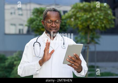 An African-American male doctor is standing near the hospital, holding a tablet in his hands. Talks to the camera on a video call with the patient. He advises, greets with his hand, reassures. Stock Photo