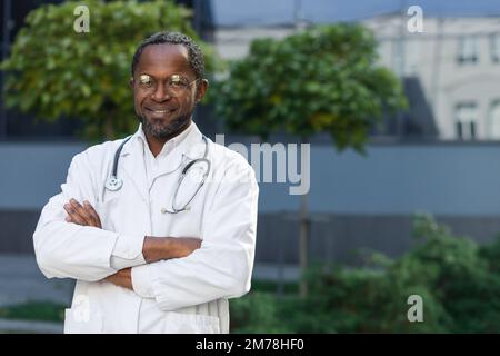 Portrait of a confident and successful young African American male doctor. He stands outside the hospital in a white coat, glasses, and a stethoscope. He looks at the camera, crosses his arms, smiles. Stock Photo