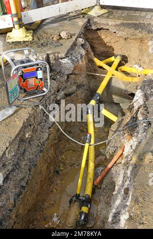 Gas main replacement pit in residential pavement new yellow plastic gas main tubes entering old ageing steel pipes and onwards to each home England UK Stock Photo