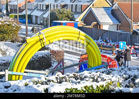 Winter snow mother pushes pram through shut down icy road works in residential village street passing large coiled up yellow gas main pipe England UK Stock Photo