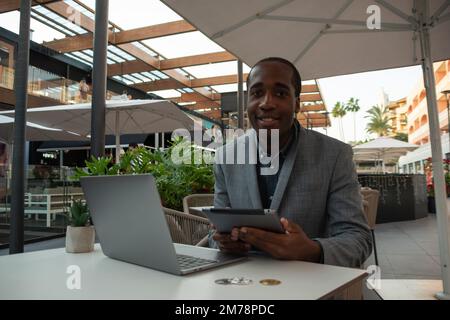 Young African trader monitors the market with laptop and tablet; the subject smiles at the camera Stock Photo