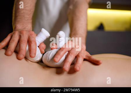 Close-up view of male masseur's hands putting herbal bags on female back during Thai massage Stock Photo