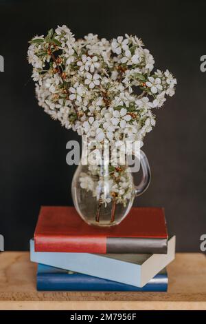 Glass vase with apple tree spring flowers sitting on a stack of books, placed on a wooden table, isolated on black background. Home decoration Stock Photo