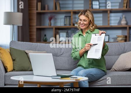 Portrait of a woman at home teaching online remotely, the teacher sits on the sofa and shows a document to the camera of the laptop online classes. Stock Photo