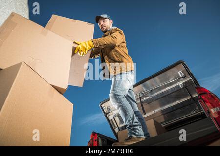 Moving Company Worker Loading Big Cardboard Boxes on the Cargo Bed of His Pickup Truck. Relocation Services Theme. Stock Photo