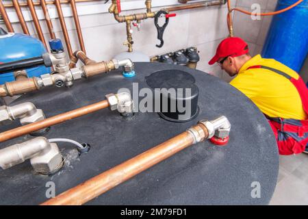 Aerial View of Tank-Type Hot Water Heater Inside Residential Boiler Room. Plumber Performing Heating System Maintenance in the Background. Stock Photo