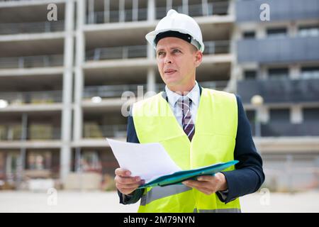 Foreman in protective helmet and vest with folder of documents on construction site Stock Photo