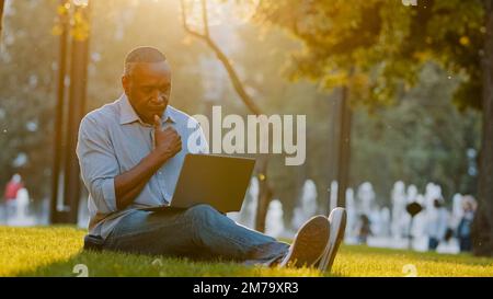 Elderly adult freelancer author writer journalist or blogger african american man sitting outdoors on grass using laptop wireless Internet on modern Stock Photo