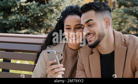 Hispanic couple sit on bench in autumn park young guy hold phone man and girl talk look at screen device surprised family smiling using app make Stock Photo