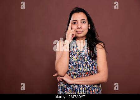 Smiling thoughtful indian woman posing portrait, model holding finger on face. Pensive person standing, looking at camera, front view studio medium shot on brown background Stock Photo