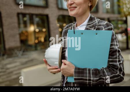 Close up of female engineer holding protective helmet and tablet standing on building background Stock Photo