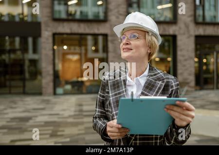 Smiling female engineer in suit with white safety helmet holding tablet on city building background Stock Photo
