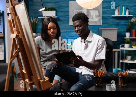 Young african american artistic partners watching video tutorials of drawing techniques on electronic tablet sitting in home art workshop. Friends having good time learning new sketching abilities. Stock Photo