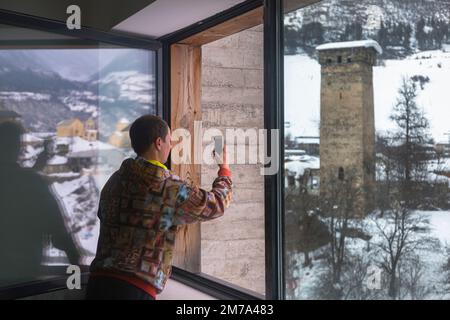 Mestia, Georgia - 2 Feb, 2020: A man takes a picture of the Svan towers from the window of the museum on his smartphone Stock Photo