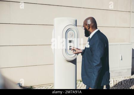 Side view of a dandy hairless African man in a tailored navy blue suit jacket checking the connection of the car charging station on the outdoors, in Stock Photo
