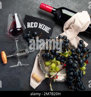 An overhead photo of glasses of red wine with a bottle, grapes, and a vintage corkscew and corks, shot from above on a dark background. Top view. Stock Photo
